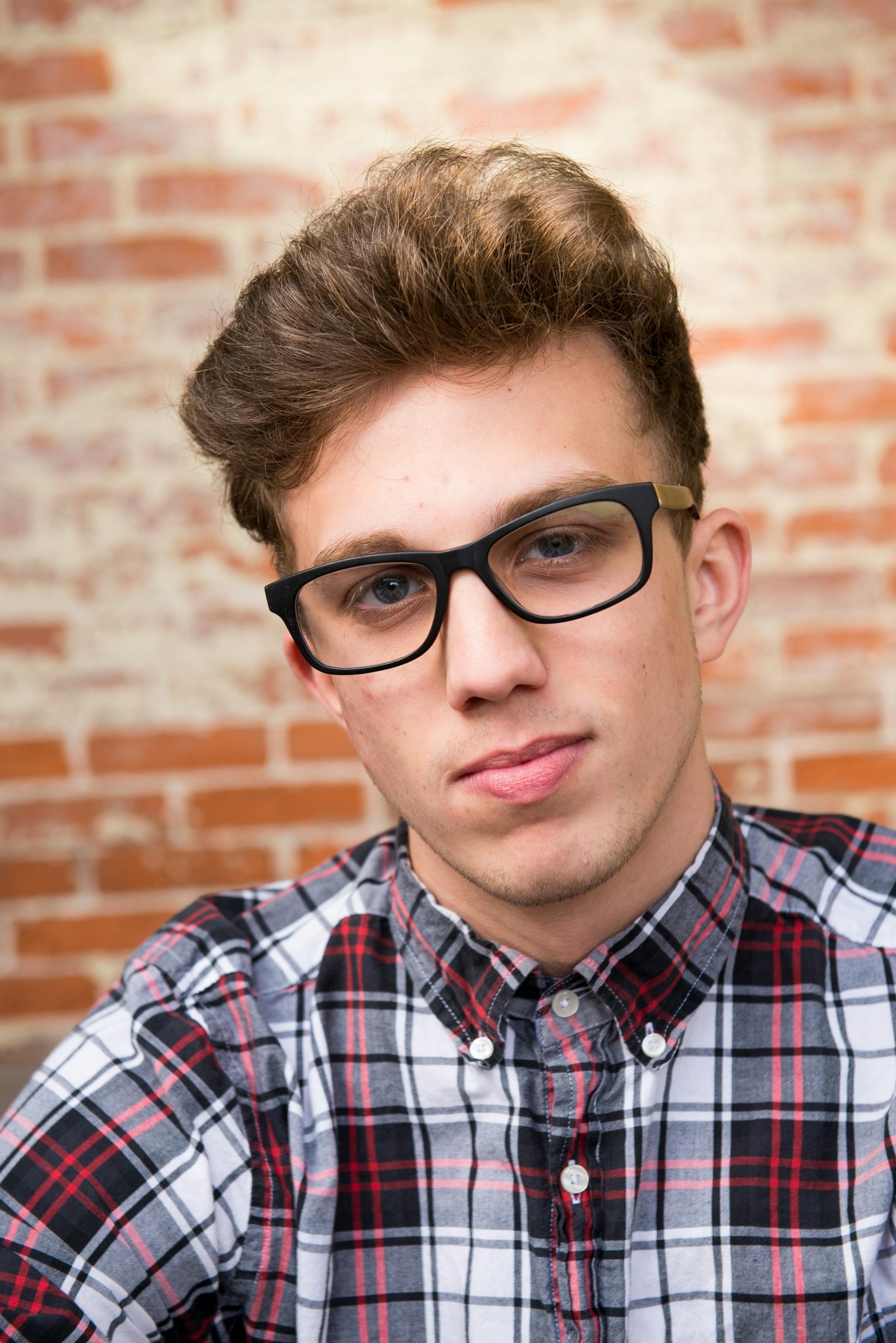 Portrait of a young man wearing eyeglasses and a plaid shirt against a brick wall.